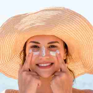 smiling woman with hat is applying sunscreen on her face